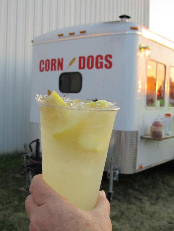 A hand holding up a glass of lemonade with a food truck that says "Corn Dogs" in the background