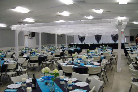 Inside of the 4-H / FFA Rental building with circular tables decorated with yellow and white flowers, blue table runners, and white candles with blue ribbon.