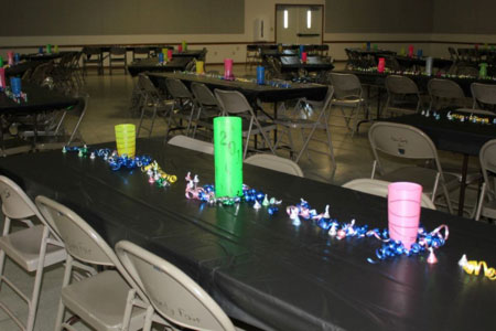 Inside of the 4-H / FFA Rental Building with rectanble tables with black tablecloths, blue, pink, and yellow decorative ribbons, and yellow, red, blue and green cylinder decorations.
